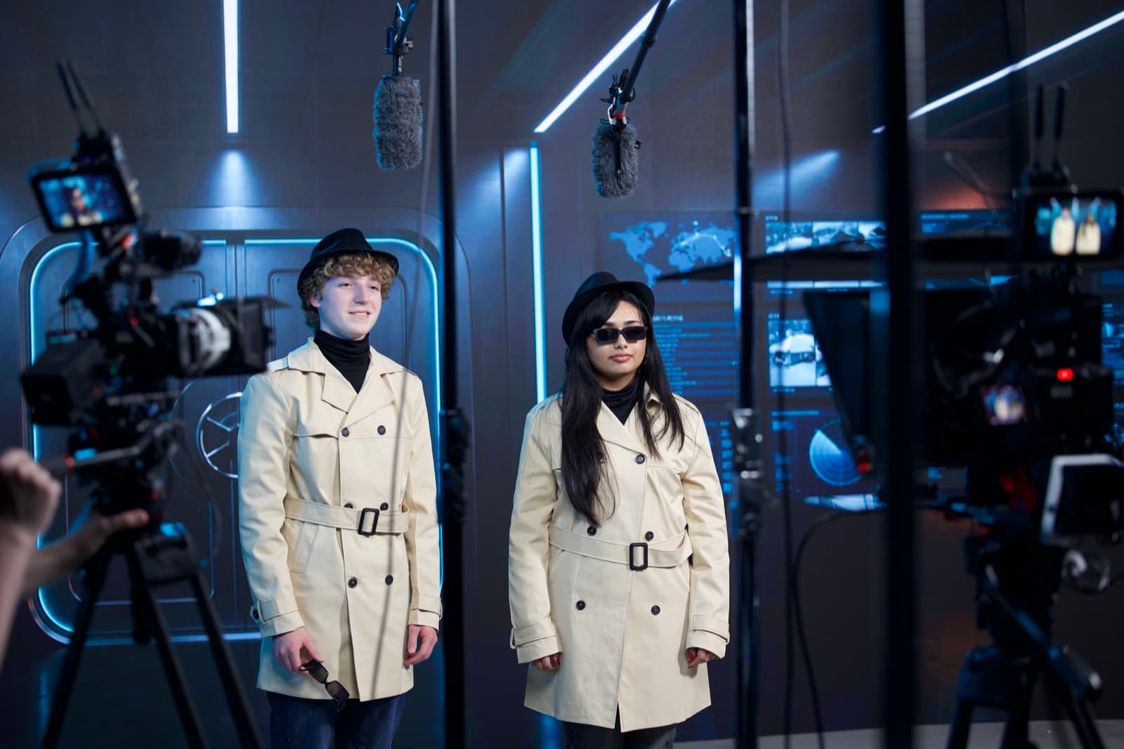 Two cinema cameras framing two on-camera hosts on a lit set, with boom microphones overhead