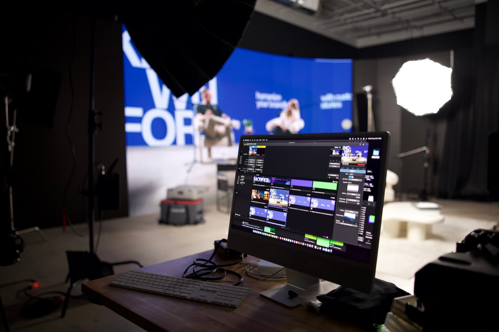 An editing timeline is open on a desktop computer in the foreground while a studio set with a blue LED wall sits out of focus behind it
