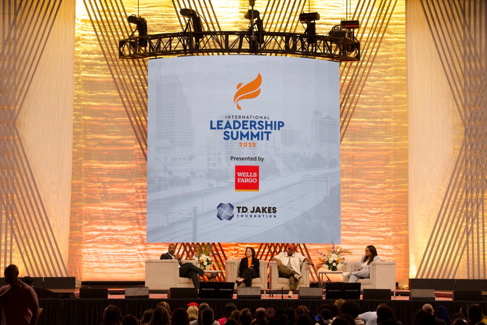 Four panelists in armchairs speak on a large stage beneath a suspended screen and circular lighting truss with a warm draped backdrop