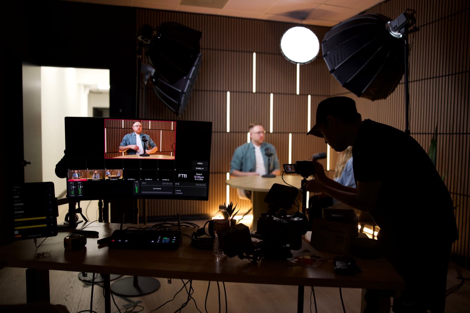 A crew member adjusts a camera beside a multiview monitor showing six camera feeds while two people are recorded at a round table under studio lights