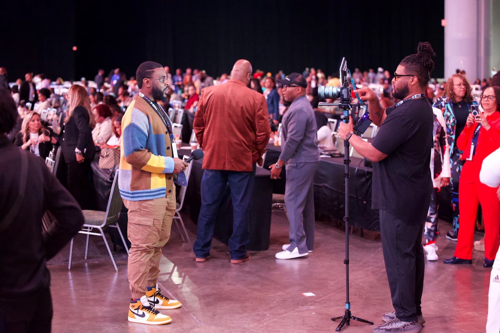 A crew member steadies a monopod-mounted camera to film an on-camera interview on a busy conference floor with attendees in the background
