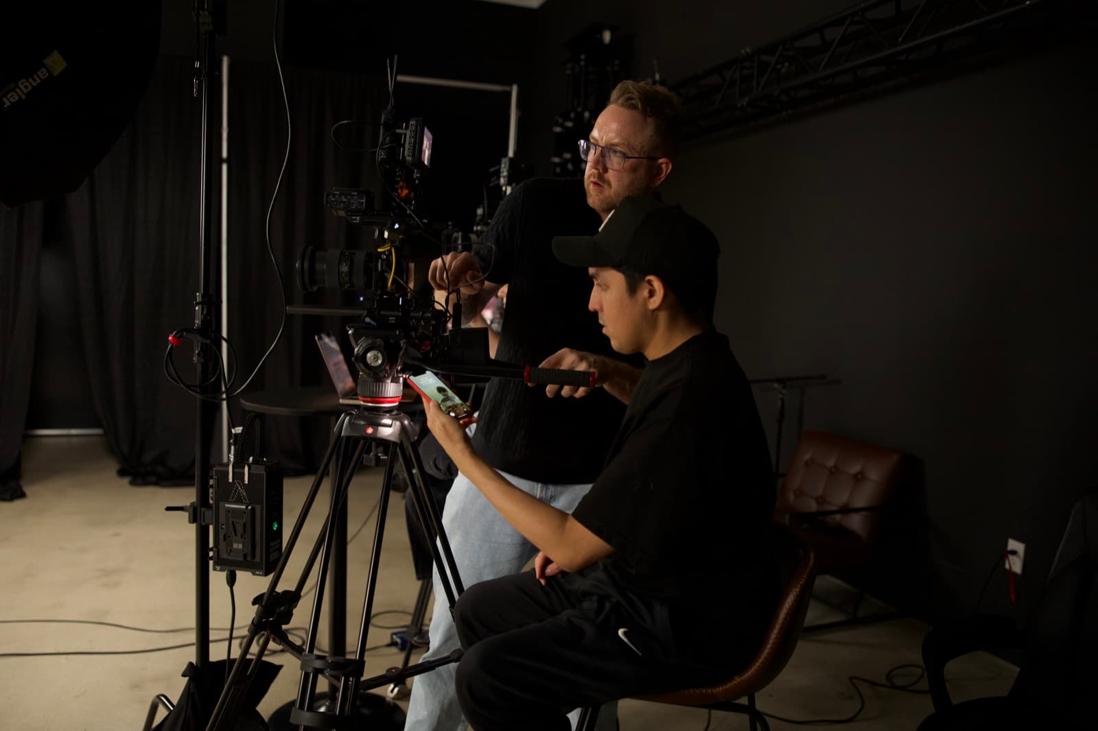 Two crew members work together at a tripod-mounted cinema camera in a studio, one adjusting the rig while the other reviews a shot
