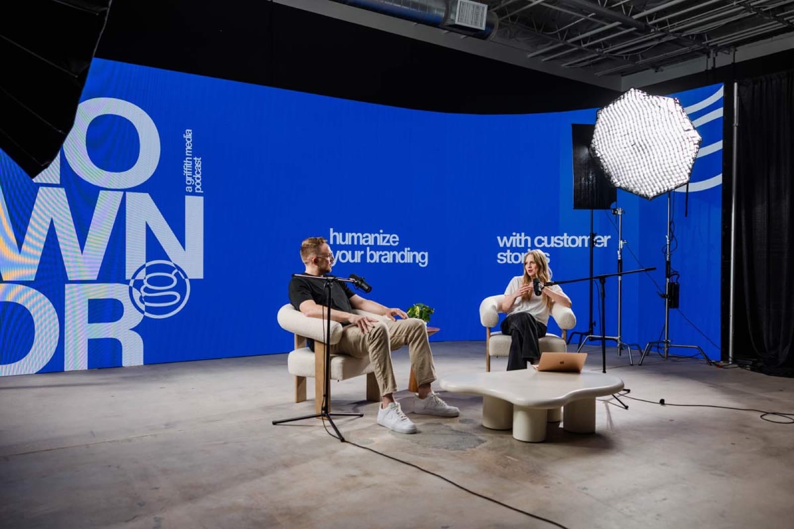Two hosts talk into boom-mounted microphones from armchairs on a podcast set in front of a large blue LED wall, lit by an octagonal softbox