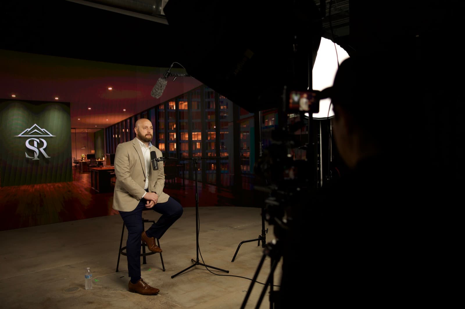 A man in a blazer sits on a stool under a soft key light with a microphone in front of him while a camera operator frames the shot