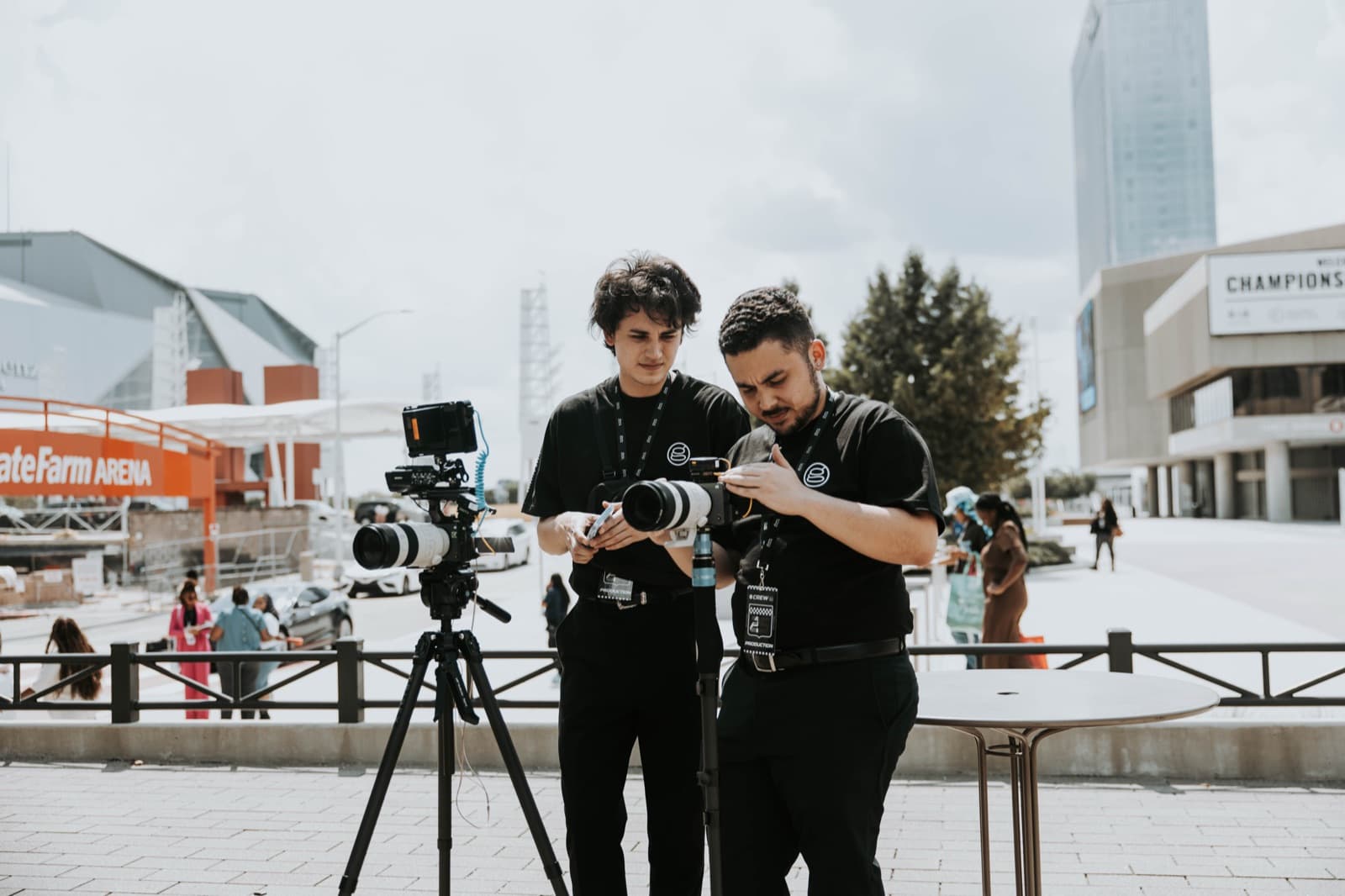 Two camera operators check cameras with long lenses on tripods on an outdoor plaza outside an arena on a bright day
