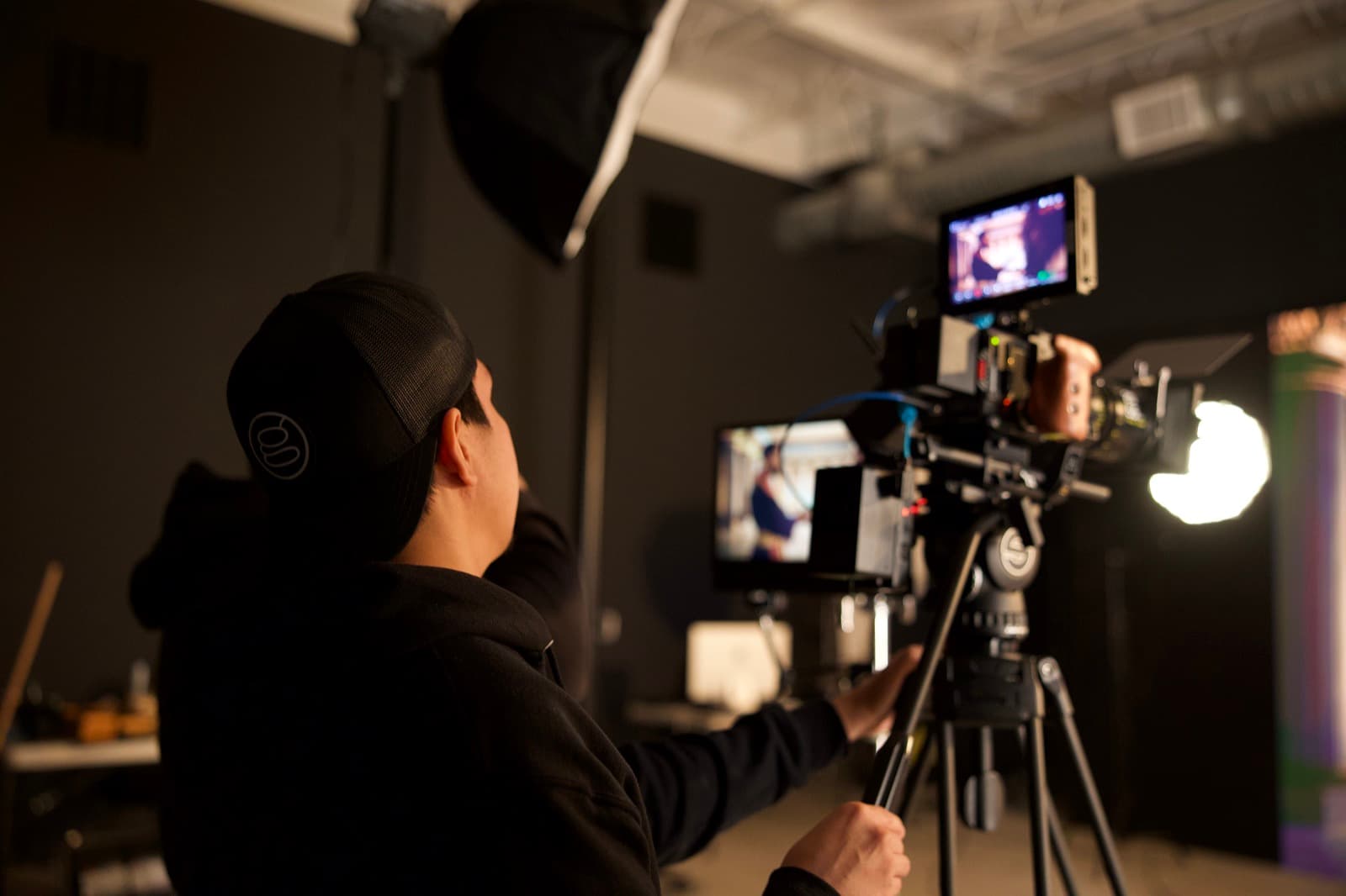 A camera operator lines up a shot on a tripod-mounted cinema camera in a dark studio, with a softbox overhead and playback monitors glowing nearby