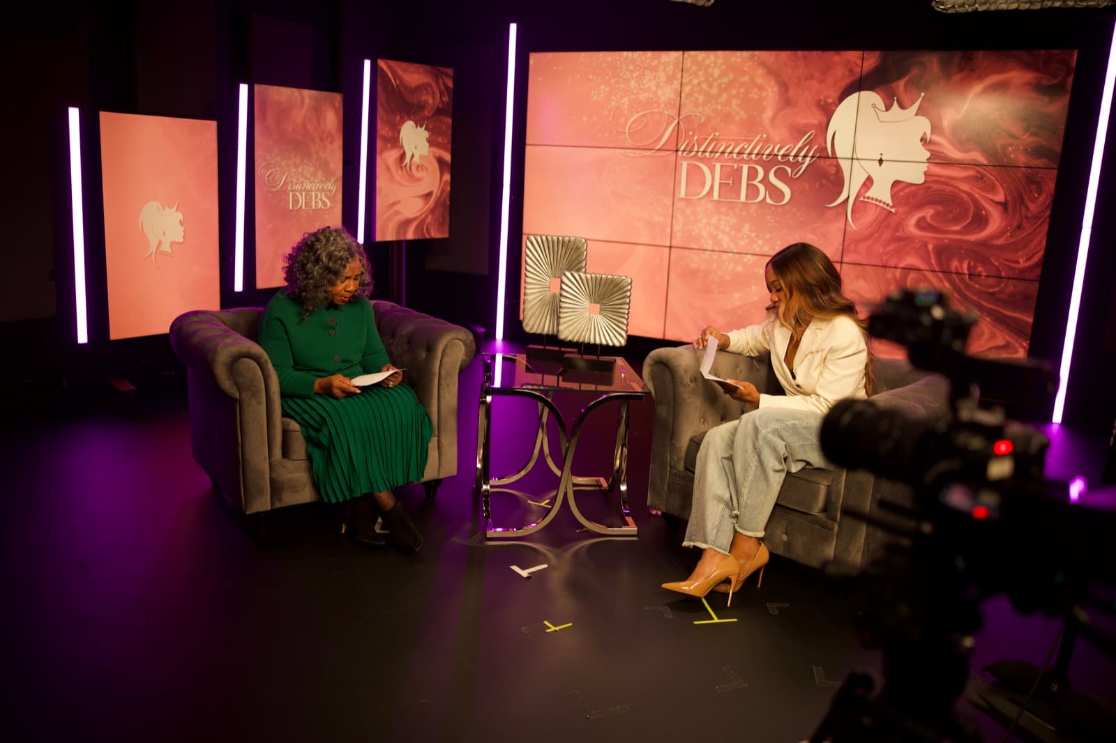 A wide studio set with two people seated for a taping between LED panel backdrops, accent lighting, and a cinema camera in the foreground