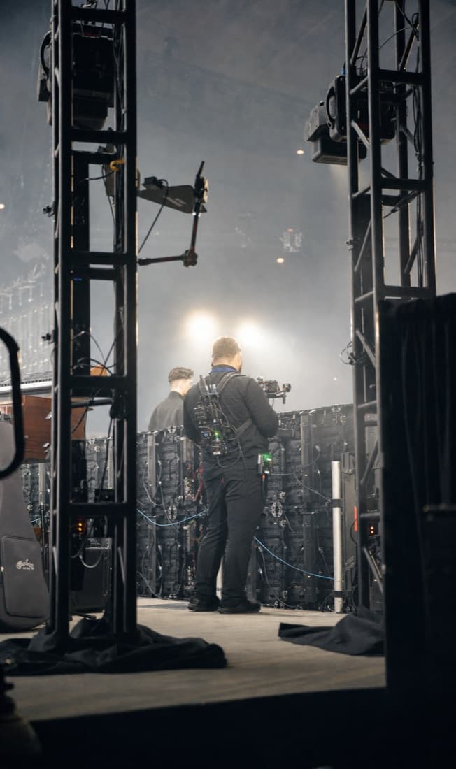 A Griffith camera operator with a wireless cinema rig backstage between truss towers, LED panels behind him.