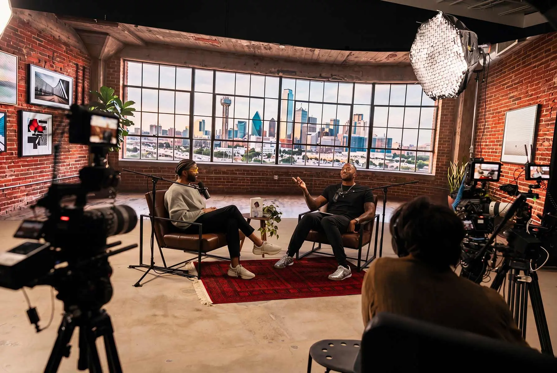 A taping on the Big Screen Stage: two hosts seated in front of the LED wall showing the Dallas skyline, surrounded by cameras and lighting.