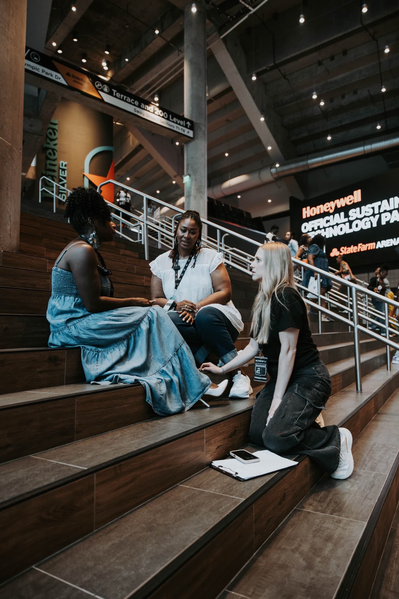 A Griffith Media crew member kneeling with a clipboard, talking with two attendees on the arena stairs