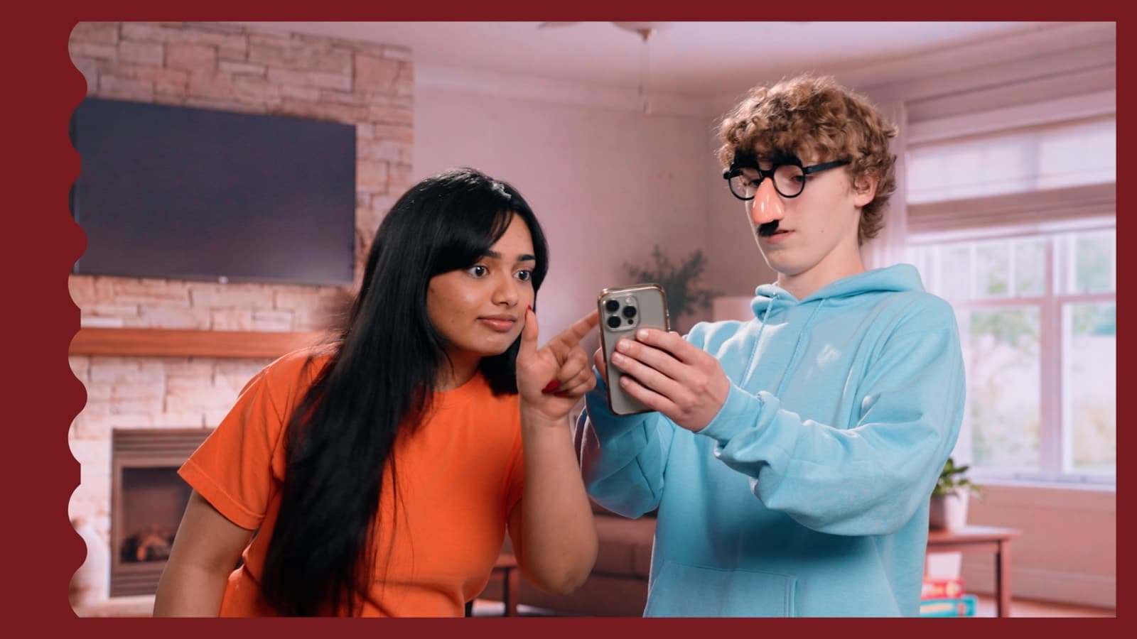 On set: a teenage actor in novelty disguise glasses and nose shows his phone to a second young actor, who points to her own cheek, mid-take in a living room set